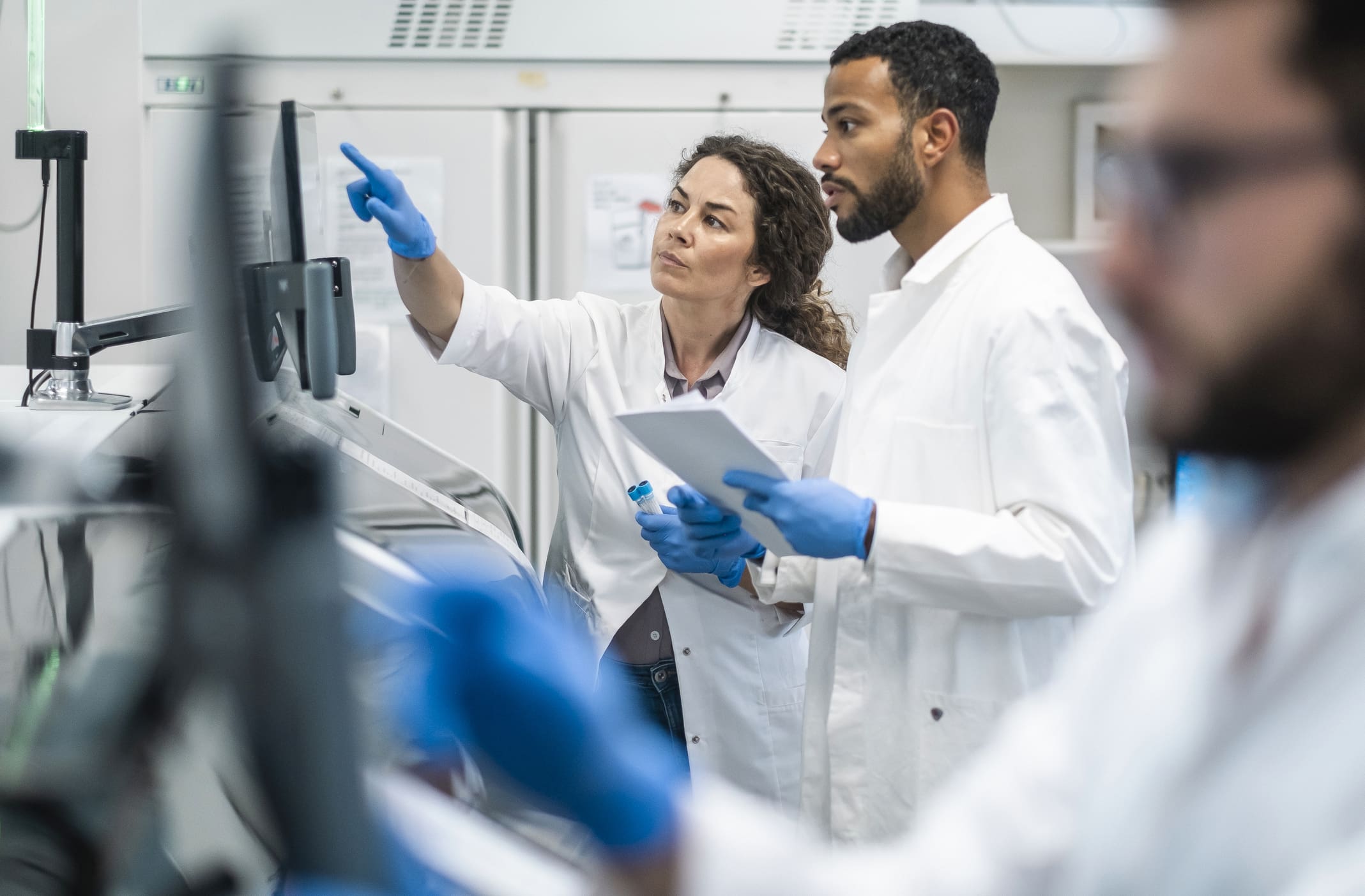 A mid-adult Caucasian woman and a mixed race man, both clad in lab coats, engage intently over research data in a bright laboratory setting.